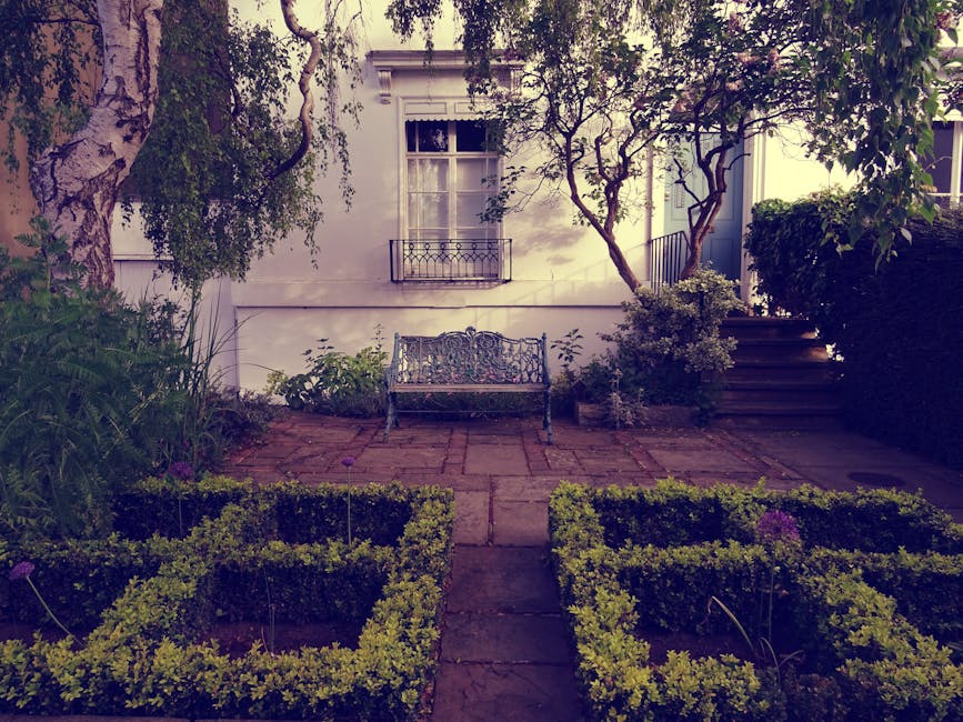 A small, well-maintained front garden in Russell Square featuring a paved pathway leading to a decorative metal garden bench positioned against a white exterior wall of a building. The garden is borde