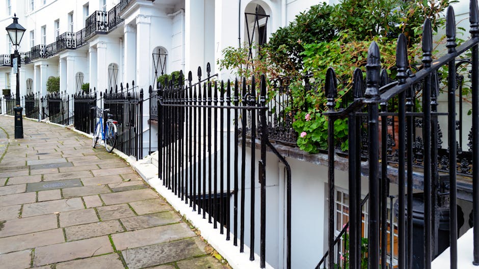 A paved pathway composed of irregularly shaped stone slabs runs alongside a white building in a residential area. A black wrought iron fence with pointed finials borders the edge of the pathway, separ